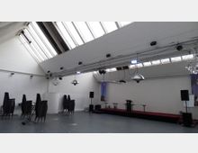 Großer, heller Veranstaltungsraum mit schrägen Decken und einer Reihe von Oberlichtern. Im Raum befinden sich gestapelte Stühle, ein Podium mit Mikrofonen sowie Lautsprecher, und die Wände sind schlicht weiß gestrichen.