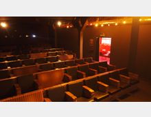 Ein kleiner, gemütlicher Kinosaal mit gestreiften Polstern auf den Sitzlehnen und gepolsterten Armlehnen aus Backsteinoptik. Im Hintergrund ist eine geöffnete Tür zu sehen, durch die ein hell beleuchteter Vorraum erscheint.