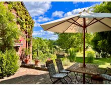 Garten mit gepflasterter Terrasse neben einer mit Efeu bewachsenen Backsteinwand, möbliert mit einem Holztisch, Stühlen und einem großen Sonnenschirm. Im Hintergrund grüne Wiesen, Büsche und Bäume unter einem blauen Himmel mit einigen Wolken.