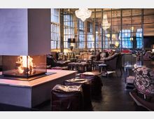 Gemütliche Lounge in einem industriell gestalteten Raum mit großen Fenstern, einem zentralen, modernen Kamin, Ledermöbeln und dekorativen Kronleuchtern, die eine warme Atmosphäre schaffen.