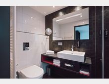 Modernes Badezimmer mit weißen Wandfliesen und schwarzer Akzentwand aus kleinen Kacheln. Zu sehen sind ein Waschbecken mit großem Spiegel, ein Wand-WC, ein Handtuchheizkörper und offene Regalfächer mit Handtüchern.