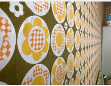 Nahaufnahme einer Wand mit Retro-Tapete in einem gelb-orange-grünen Blumenmuster auf olivgrünem Hintergrund. Rechts im Bild sieht man unscharf ein Regal mit Gegenständen.