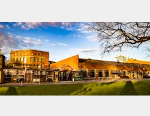 Backstein-Gebäudeensemble mit industriellem Charme, bestehend aus einem großen, flachen Bau mit der Aufschrift „Festsaal Kreuzberg“ und einem mehrstöckigen historischen Gebäude im Hintergrund. Der Vordergrund zeigt eine Rasenfläche, gepflasterte Wege sowie einen Blick auf alternative Holzkonstruktionen und einen Verkaufsstand.