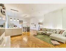 Offener Wohnraum mit Parkettboden, bestehend aus einer modernen Küche mit Kücheninsel und Barhockern sowie einem Wohnbereich mit einer großen, hellgrauen Ecksofa, Couchtisch und grünem Teppich. Helle Wände, dezente Beleuchtung und dekorative Elemente wie Regale und Kunstwerke prägen die Umgebung.