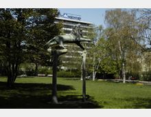 Bronzestatue eines springenden Löwen, gestützt von zwei Säulen, in einem Park mit grüner Wiese und Bäumen; im Hintergrund ein modernes Bürogebäude mit "HOCHTIEF"-Schriftzug.