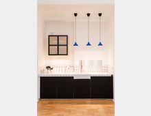 Moderne Küchenzeile mit schwarzen Unterschränken und weißer Arbeitsplatte, gefliest mit kleinen weißen Kacheln. Darüber hängen drei blaue Pendelleuchten, und an der Wand befindet sich ein hängender Wandschrank mit Gittertüren. Der Boden besteht aus hellem Holzparkett.