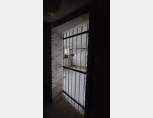 Ein dunkler Kellerraum mit freiliegenden Ziegelwänden und einer Metallgittertür im Vordergrund. Im Hintergrund ist ein kleines Fenster zu sehen, das etwas Licht in den Raum bringt.