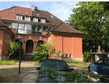 Ein zweistöckiges Haus mit roter Fassade und dunkelbraunem Ziegeldach. Vor dem Gebäude befinden sich drei Rundbögen im Eingangsbereich, umgeben von einem kleinen, grünen Garten mit Bäumen und Sträuchern. Zwei geparkte Autos sind im Vordergrund zu sehen.