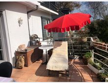 Möblierte Terrasse mit Terrakottafliesen, einem roten Sonnenschirm, einer Liege mit heller Polsterung und verschiedenen Blumentöpfen. Im Hintergrund ist eine weiße Hauswand mit Fensterläden und Geländer zu sehen; umgeben von Bäumen und Pflanzen.