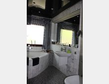 Kleines Badezimmer mit Marmorelementen an Wänden und Badewanne, einer abgerundeten Badewanne in der Ecke, einem Waschbecken und einem Spiegel. Der Raum hat ein Fenster mit gemusterten Vorhängen und einen Boden aus kleinen Mosaikfliesen.