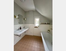 Helles Badezimmer mit schräger Dachdecke, weißen Wandfliesen und beigefarbenem Fliesenboden mit sechseckigem Muster. Links ein Doppelwaschbecken mit Spiegel und Wandleuchten, rechts eine Badewanne; mittig ein kleines Fenster über einer schmalen Ablage.