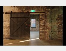 Industriehalle mit freigelegtem Ziegelmauerwerk und einer großen hölzernen Schiebetür. Durch die geöffnete Tür ist ein weiterer Raum mit einem Fenster im industriellen Stil sichtbar, beleuchtet von Tageslicht.