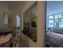 Ein Innenraum mit mehreren Bereichen: Im Vordergrund eine weiße Wand mit einer Glaselement-Einlage aus runden, strukturierten Glasscheiben. Links eine halb geöffnete, geschwungene Tür, die zu einem anderen Raum führt. Rechts ein Wohnzimmer mit großen Fenstern, einem Heizkörper, einem Teppich und einer grauen Couch.