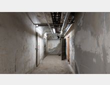 Ein schmaler, verlassener Flur mit abgenutzten, grauen Wänden, teilweise sichtbaren Rohren und Kabeln an der Decke sowie mehreren Metall- und Holztüren, teils geöffnet. Der Boden ist aus Beton und weist Verschmutzungen auf.