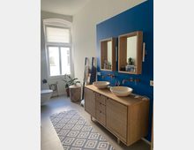 Modernes Badezimmer mit einer Wand in kräftigem Blau, einem Holzwaschtisch mit zwei Aufsatzwaschbecken und kupferfarbenen Armaturen. Links ein großes Fenster mit halb geschlossenem Rollo, ein WC, ein Teppich mit geometrischem Muster sowie Pflanzen und ein Wäschekorb.