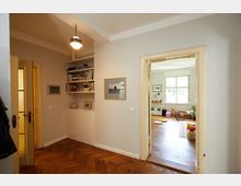 Flur einer Wohnung mit Holzboden und hellgrauen Wänden, die in ein weiteres Zimmer führen. An der linken Wand befindet sich ein eingebautes Regal mit Büchern und Dekorationen, der Raum im Hintergrund hat ein großes Fenster, Spielzeug und einen grünen Sessel.