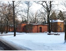 Backsteingebäude in einer winterlichen Parklandschaft mit schneebedecktem Boden, kahlen Bäumen und Sitzbänken im Vordergrund. Das Hauptgebäude ist einstöckig mit einem gewölbten Eingang, daneben steht ein zweistöckiges Gebäude mit Satteldach und großen Fenstern.