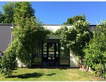 A modern building with dark-framed glass doors and windows, surrounded by lush green plants and flowering shrubs. Behind the building, tall trees and a clear blue sky are visible, with a well-maintained grassy lawn in the foreground.