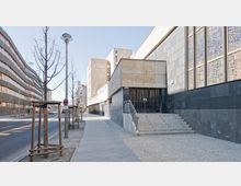 Straßenszene mit modernen Gebäuden: Rechts ist ein Gebäude mit dunkler Fassade und großen Fenstern mit abstraktem Muster zu sehen, davor eine Treppe zum Eingang. Links verläuft eine Bürgersteig- und Straßenfläche mit jungen, gestützten Bäumen und weiteren modernen Gebäuden im Hintergrund.