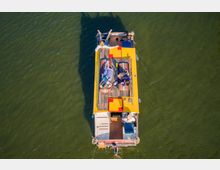 Luftaufnahme eines kleinen Motorbootes auf grünem Wasser, auf dem drei Personen auf Liegeflächen im Sonnendeckbereich entspannen; das Boot hat ein gelbes Dach und Holzboden, und im Heckbereich sitzt eine Person am Steuerrad.