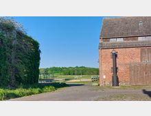 Backsteingebäude mit Holzfenstern und Schornstein auf der rechten Seite, daneben eine mit Efeu bewachsene Wand, im Hintergrund eine grüne Wiese mit Holzumzäunung unter blauem Himmel.