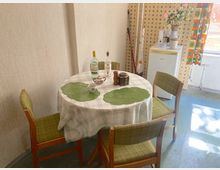 Ein kleiner Essbereich mit einem runden Tisch, gedeckt mit einer karierten Tischdecke und grünen Platzdeckchen, umgeben von vier Holzstühlen mit grünen Polstern. Im Hintergrund steht ein weißer Kühlschrank neben einer Wand mit orange-gelber Retro-Tapete und einem Fenster mit buntem Vorhang.