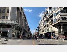 Straßenszene in einer Stadt mit modernen Gebäuden auf beiden Seiten, links ein rechteckiges Gebäude mit Glasdach und rechts ein Gebäude mit schrägen, gestaffelten Fassaden und vielen Fenstern; in der Mitte eine Straße mit Radfahrern und Fußgängern unter blauem Himmel.