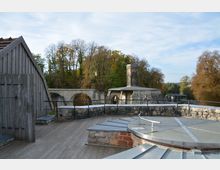 Historische Industrieanlage mit gemauerten Steinstrukturen, darunter ein runder Schornstein und Bogengänge. Im Vordergrund eine Terrasse mit Holzboden, Metallgeländern und einer Abdeckung, umgeben von herbstlicher Baumlandschaft.