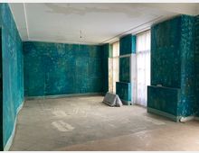 Ein leerer Raum mit türkisfarbenen, abgenutzten Wänden und einem fleckigen, hellen Boden. Große Fenster mit weißen Vorhängen lassen Tageslicht herein, während an einer Wand eine freistehende Struktur oder Abdeckung zu sehen ist.