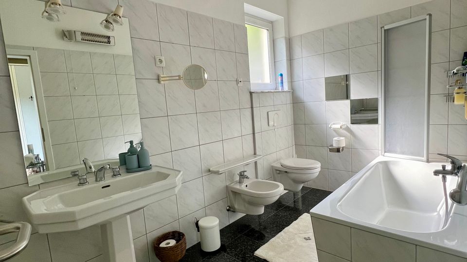 Helles, gefliestes Badezimmer mit großem Waschbecken und Spiegel, Bidet und Toilette sowie einer Badewanne mit Duschabtrennung. Dunkler Steinboden, weiße Badvorleger und ein kleines Fenster sorgen für Kontrast und Tageslicht.