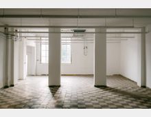 Leerer Innenraum mit weißen Wänden und großen Fenstern, strukturiert durch breite Säulen. Der Boden ist mit schwarz-weißen Schachbrettfliesen ausgelegt, und an der Decke sind Rohre und Leuchtstofflampen angebracht.