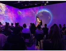 Eine Person spielt Harfe vor einem sitzenden Publikum in einem abgedunkelten Raum mit großen Projektionen auf den Wänden, die eine animierte Weltraumszene mit Planeten und einem leuchtenden Globus zeigen. Die Beleuchtung ist violett und atmosphärisch.