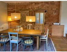 Esszimmer mit rustikalem Stil, bestehend aus einem Holztisch, umgeben von verschiedenen Stühlen. Die Rückwand besteht aus unverputztem roten Backstein mit einer Holztür in der Mitte. Zwei hängende Lampenschirme beleuchten den Raum, der auf einem Teppich mit Holzfußboden liegt.