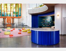 Moderne Kircheninnenraum mit farbigen Elementen: Im Vordergrund ein runder, blauer Tisch; auf einem rosafarbenen Teppich liegen bunte, runde Sitzkissen. Im Hintergrund ein orangegerahmtes Fenster mit buntem Glas und ein Altarbereich; rechts eine blaue Wand mit Regalen, auf denen Bücher und kleine Objekte ausgestellt sind.