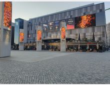 Großer, moderner Platz mit gepflastertem Boden vor einem würfelförmigen Gebäude mit grauen Fassadenelementen und großflächigen Werbebildschirmen. Vor dem Gebäude stehen vier schmale Säulen mit digitalen Anzeigen, im Erdgeschoss befinden sich Geschäfte und Restaurants mit Sitzbereichen im Freien.