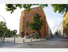 Straßenecke in einer Stadt mit einem orangefarbenen, mehrstöckigen Gebäude im Fokus, das Geschäftsräume und Wohnungen beherbergt. Im Vordergrund befinden sich ein Gehweg, Verkehrsampeln und ein Baum, während rechts und links weitere Gebäudezeilen die Straße säumen.