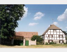 Fachwerkhaus mit Satteldach und weißen Wänden, durch schwarze Holzbalken unterteilt, neben einer Scheune mit Ziegeldach und grünen Toren, umgeben von Bäumen und einer Wiese unter blauem Himmel mit wenigen Wolken.