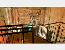 Industrieartig gestalteter Treppenabgang in einem Gebäude mit unverputzten Betonwänden und einem Mauerstück, das mit einem großen Wandbild einer stilisierten Schnecke verziert ist. Geländer aus Metall und warme Beleuchtung prägen die Szene.