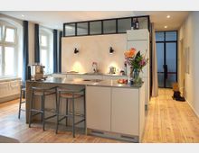 Moderne Küche mit einer zentralen Kücheninsel aus beigen Schränken und einer Arbeitsplatte aus Edelstahl, flankiert von drei Barhockern. Im Hintergrund befinden sich Fenster mit blauen Vorhängen, eine halbhohe Wand mit Glasoberlichtern sowie eine Tür mit schwarzem Metallrahmen.