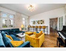 Helles Wohnzimmer mit großen Fenstern, freiliegender Putzwand und Fischgrätparkett, eingerichtet mit einer blauen Eckcouch, gelbem Sessel und Couchtisch. An der Wand steht eine schmale Bar mit hohen Stühlen, rechts neben einer Glastür ist ein Klavier zu sehen.