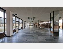 Große, leere Eingangshalle oder Foyer mit grauem Steinboden, weißer Rasterdecke und Reihen von Deckenleuchten. Entlang der linken Seite verlaufen bodentiefe Fensterfronten, rechts stehen eine Garderoben-/Thekenzone und ein Treppenaufgang neben Ziegelwänden.