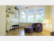 Wohnzimmer mit hellem Parkettboden und einem weißen Klavier mit schwarzem Hocker. Links sind Bücherregale, rechts befinden sich ein lila Sessel, ein passender Hocker, eine Decke, eine Lampe und große Fenster mit Blick ins Grüne.