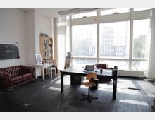 Heller Büroraum mit großen Fensterfronten, die viel Tageslicht einlassen. Der Raum ist mit zwei roten Ledersofas, einem Holztisch mit Laptop, einem Bücherregal und mehreren kleineren Einrichtungsgegenständen ausgestattet.