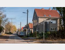 Straßenkreuzung in einem Wohngebiet mit einem großen, grauen Wohnhaus mit rotem Ziegeldach rechts im Bild. Links verläuft ein Gehweg mit geparkten Autos entlang einer mit Bäumen gesäumten Straße, im Hintergrund Stromleitungen und kleinere Häuser.