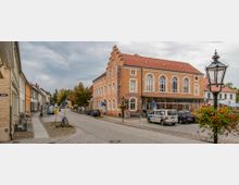 Straßenszene in einem kleinen Ort mit einem historischen Gebäude, das als Hotel und Restaurant dient. Das orangefarbene Haus hat Rundbogenfenster, ein Ziegeldach und Beschriftungen an der Fassade; die Straße ist gesäumt von weiteren mehrstöckigen Gebäuden, parkenden Autos und Laternen.