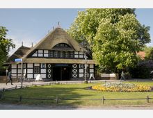Fachwerkgebäude mit Reetdach, genutzt als U-Bahn-Station „Dahlem-Dorf“, umgeben von Bäumen und Blumenbeeten. Im Vordergrund ein kleiner, kreisförmiger Blumenhof sowie ein gepflasterter Gehweg.