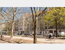 Städtischer Platz mit Bäumen und leichtem Frühlingslaub, umgeben von mehrstöckigen Wohngebäuden mit heller Fassade. Im Vordergrund fährt eine Person auf einem Fahrrad, während geparkte Autos und ein Gehweg entlang der Gebäude zu sehen sind.