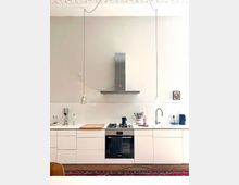 Minimalist white kitchen with a long counter and flat-front cabinets, featuring a built-in oven and gas cooktop beneath a stainless-steel range hood. Two pendant lights hang from the high ceiling, and a sink, small appliances, and utensils sit along the tiled backsplash.