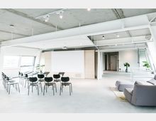 Heller, moderner Besprechungsraum mit Betonboden und freiliegender Decke, ausgestattet mit schwarzen Stühlen, einem großen weißen Präsentationsbildschirm und einzelnen Pflanzen. Große Fenster lassen Tageslicht herein, und im Vordergrund steht eine graue Couch auf einem Teppich.