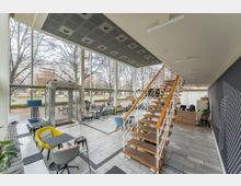 Innenansicht eines modernen Cafés mit großen bodentiefen Fenstern, die einen Blick auf Bäume und eine Straße ermöglichen. Das Café hat helle Fliesenböden, bunte Sitzmöbel, eine Holztreppe mit Metallgeländer und eine Theke im Hintergrund.